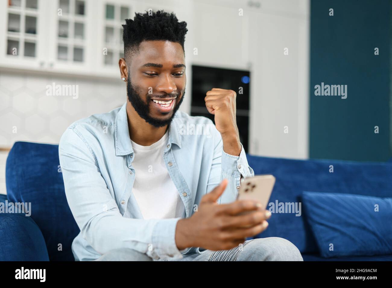 Excited black multiracial man sitting on the sofa looking at the phone and smiling, surprised emotional lucky man, rejoicing at good message, news, won the lottery Stock Photo