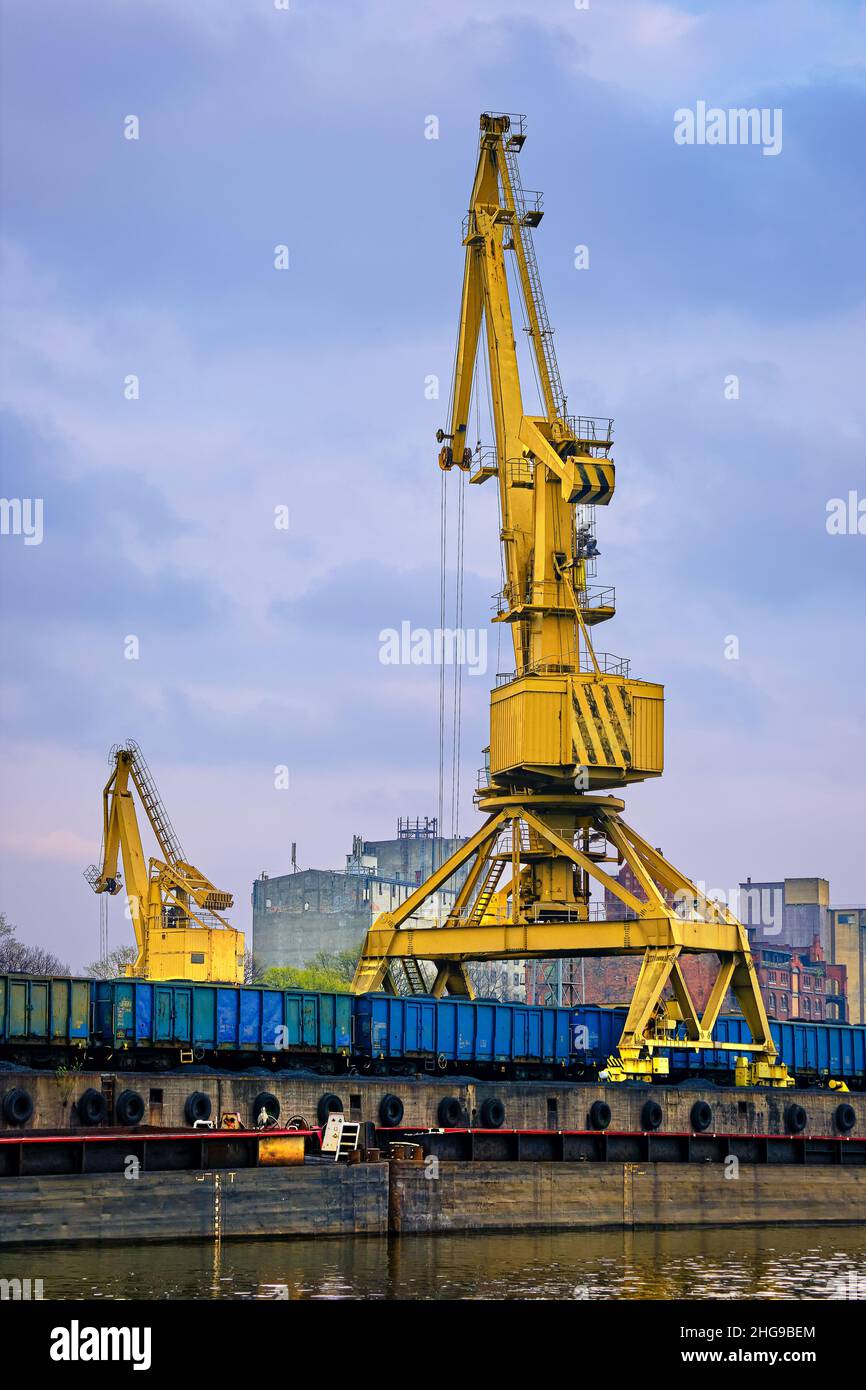River port crane loading open-top gondola cars on cloudy day. Empty river drag boats or barges moored by pier, Empty cars ready for loading. Stock Photo