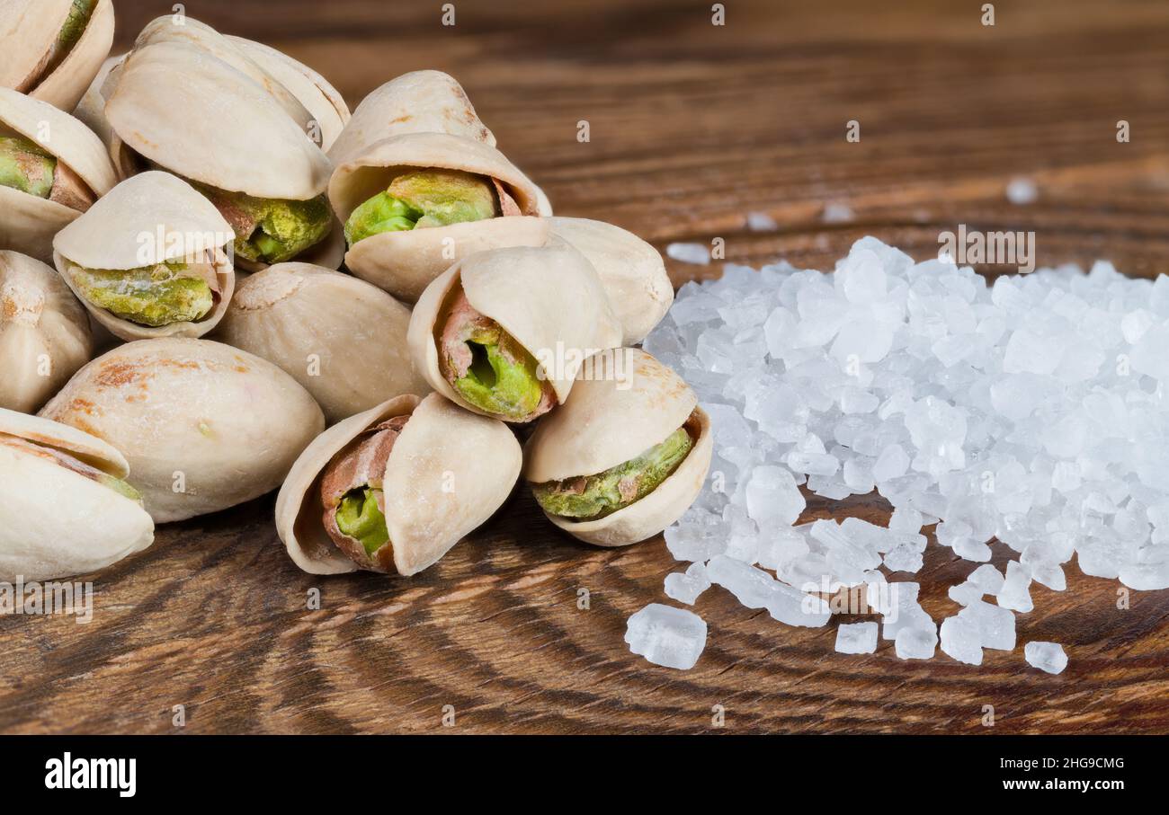 Closeup of roasted pistachio nut pile and table salt on a wooden background. Pistacia vera. Salted green fruits in open hard split halved beige shells. Stock Photo