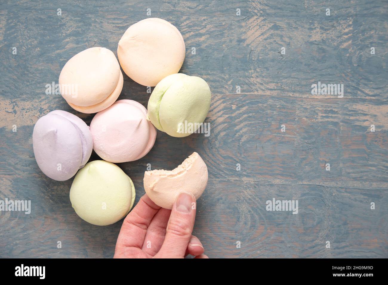 Round multi-colored marshmallows in a pile and a hand holding a bitten half of a marshmallow on a blue wooden background, top view copy space. Delicio Stock Photo