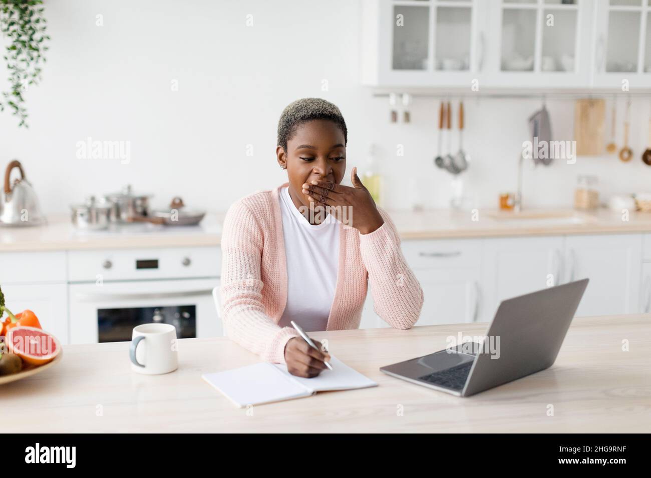 Tired young pretty black female looks at laptop makes notes, yawns at minimalist kitchen interior, free space Stock Photo