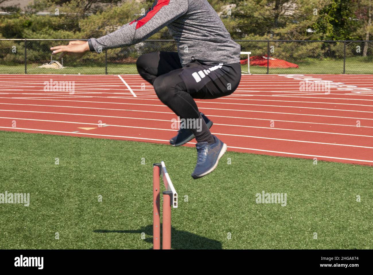 Side view of a male high school athlete jumping over a track hurdle on green truf close up. Stock Photo