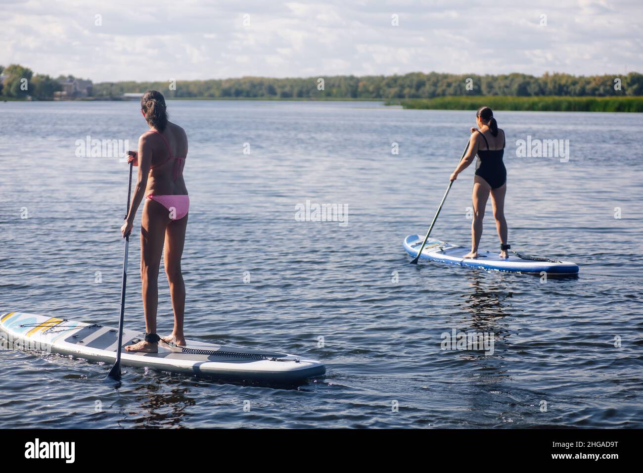 Two middle aged women puddle boarding on beautiful blue lake with oars in hands with trees and reeds in background both wearing swimming suits. Active Stock Photo