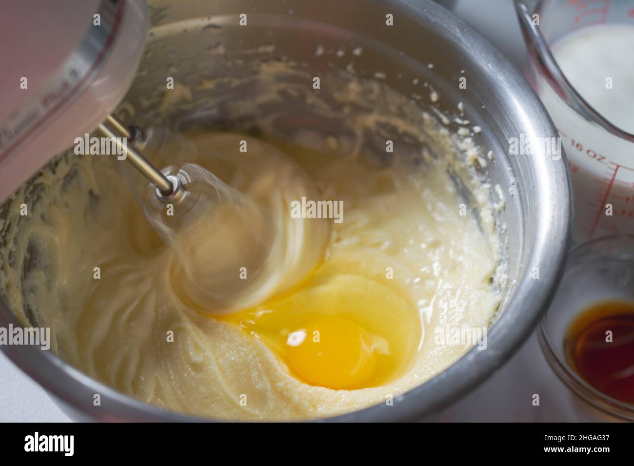 Making of a cake: Adding an egg and beating batter.  Mixer beaters showing motion blur.  Milk and vanilla in background. Stock Photo