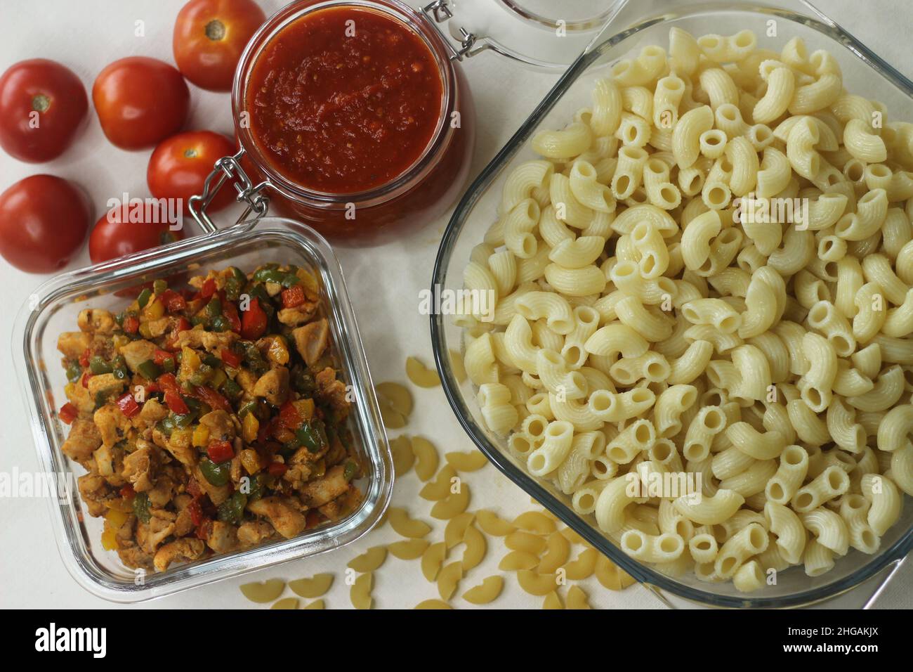 Boiled macaroni pasta inside a square glass bowl. Shot on white background with tomato red sauce in a jar, sauteed chicken and bell peppers and raw ma Stock Photo