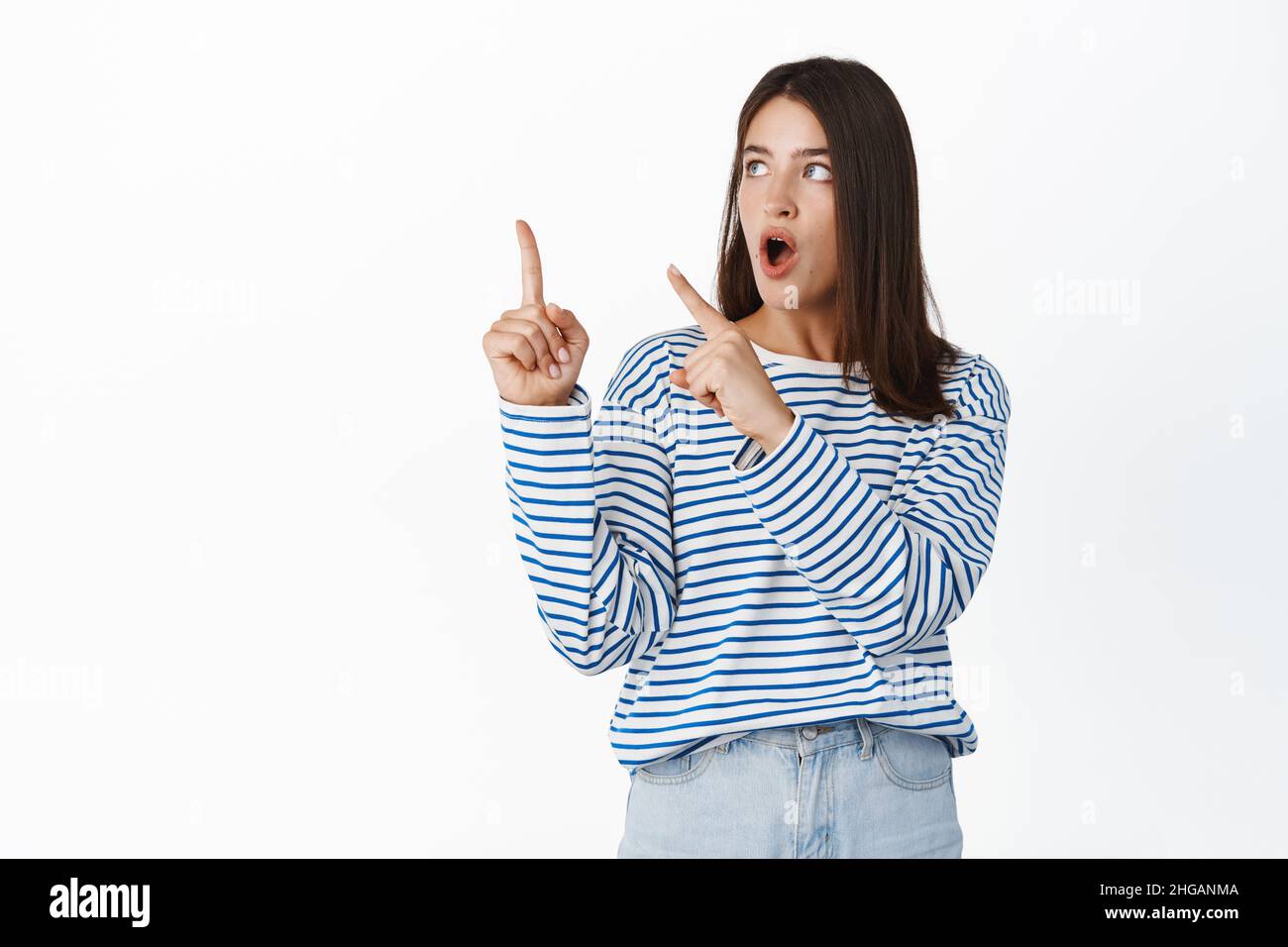 Shocked young brunette woman pointing at upper left corner with awe
