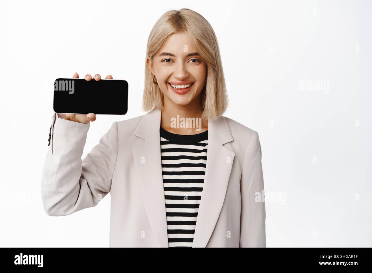 Image of smiling saleswoman showing mobile phone horizontal, demonstrating online store, company website on smartphone, standing in suit over white Stock Photo