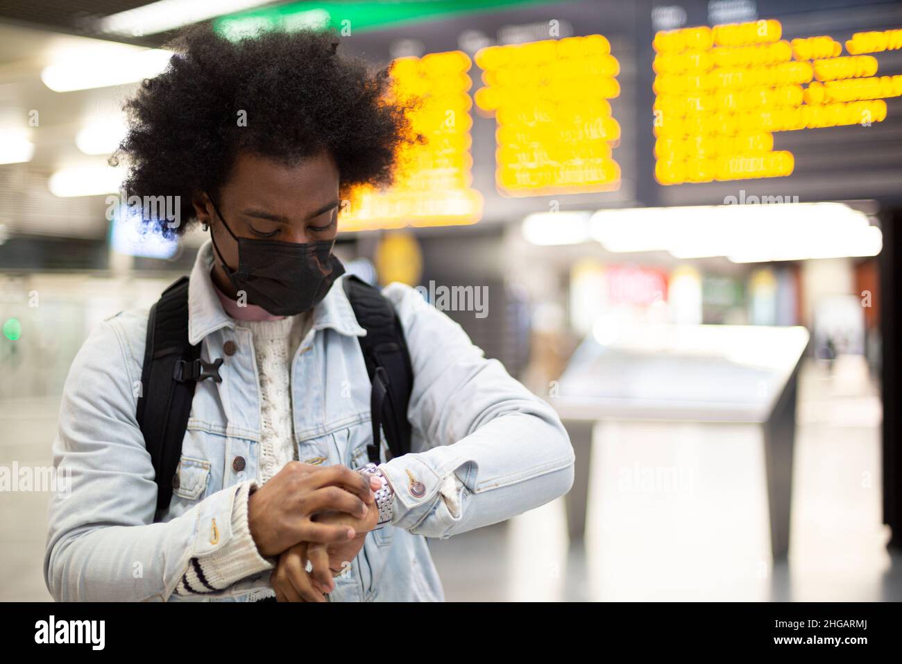 Young African American man with face mask looking at the time on his watch at the airport. Concept of delayed flights. Space for text. Stock Photo