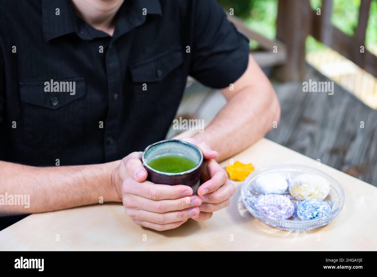 Man sitting at table with closeup of hands holding matcha green tea cup outside in backyard garden deck with Japanese daifuku mochi food Stock Photo