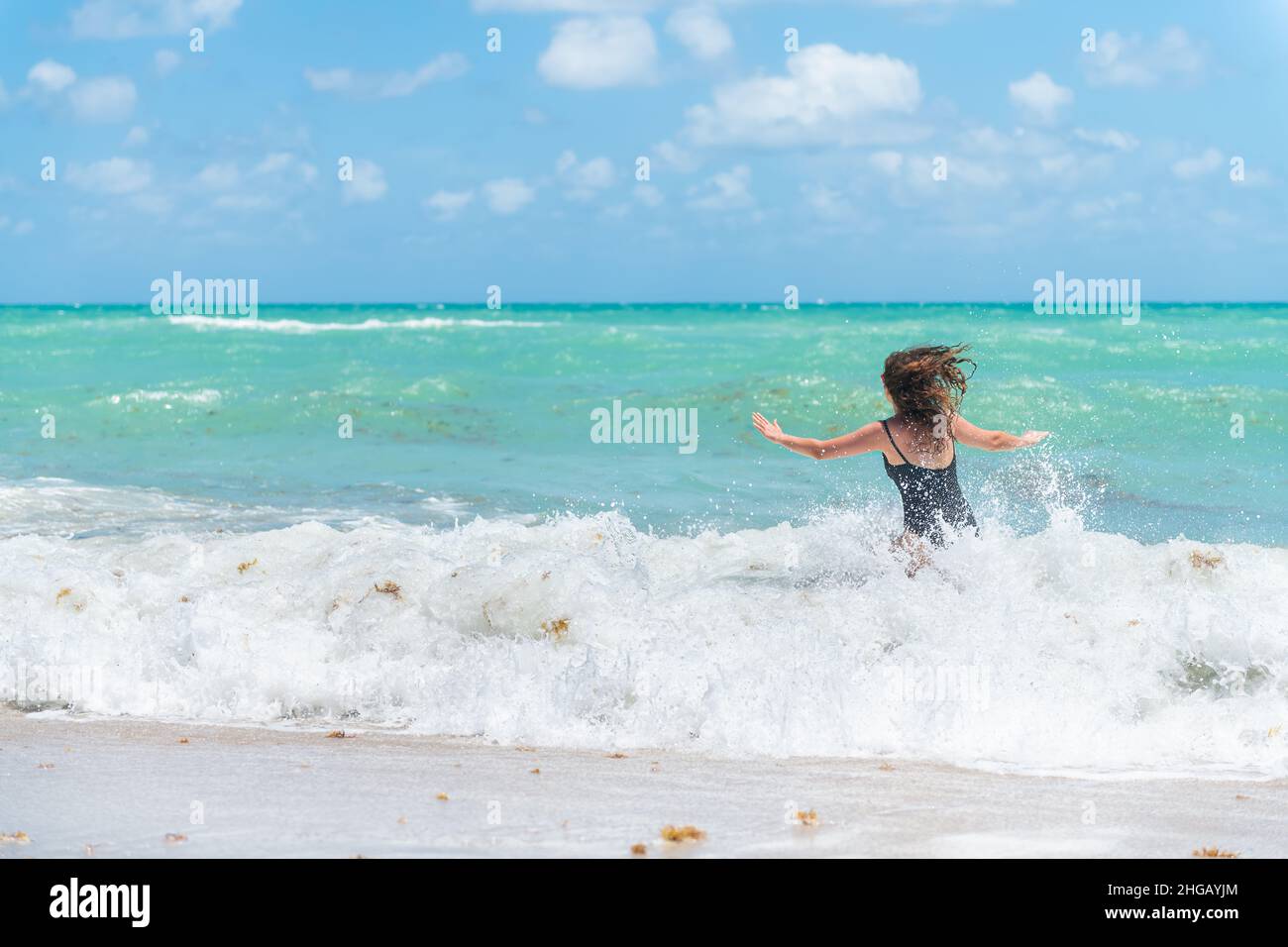 Back of young woman in swimsuit with waves splashing crashing in Sunny Isles, Miami Beach, Florida with Atlantic ocean sea green turquoise water and s Stock Photo