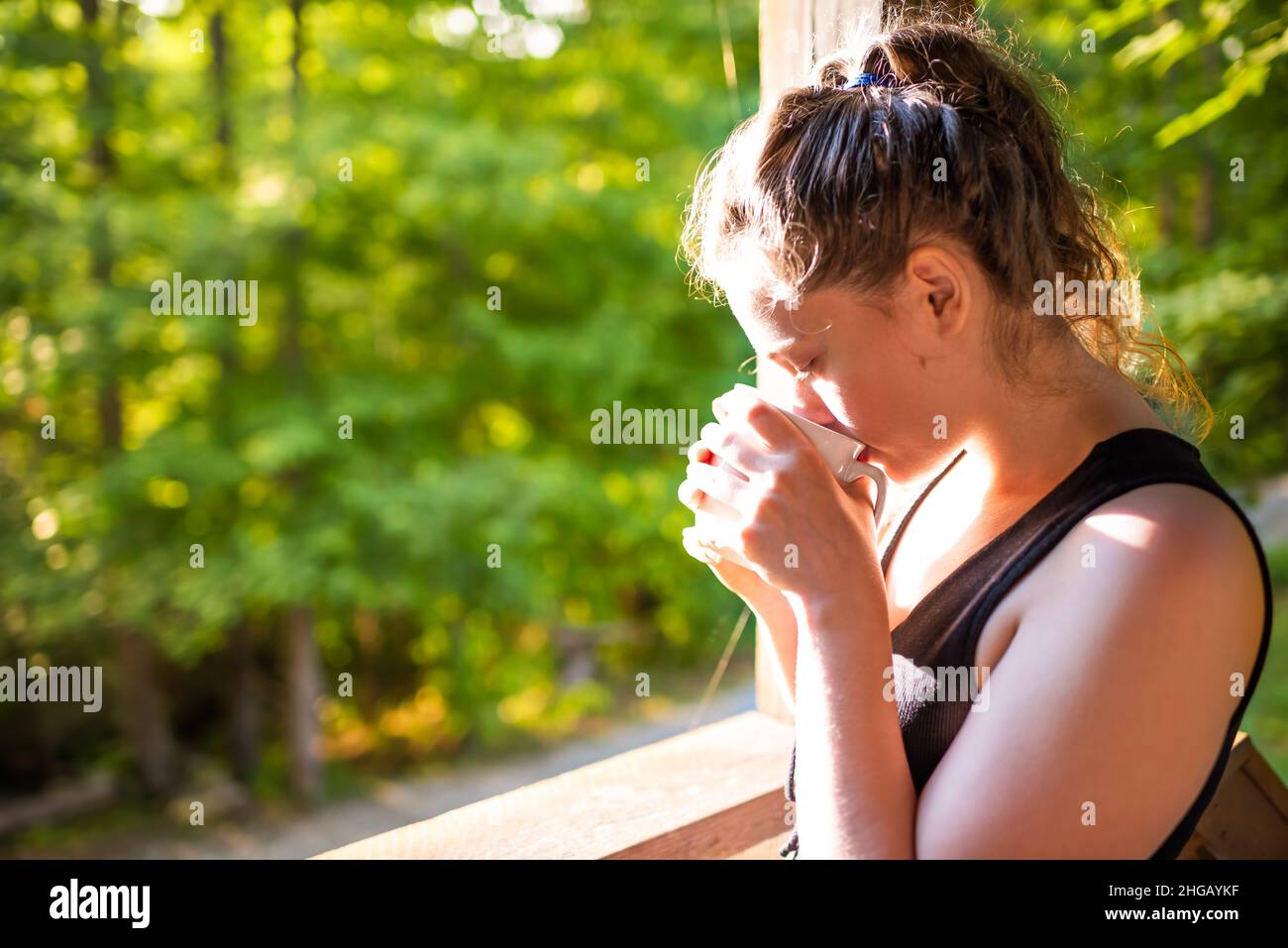 Woman drinking coffee or tea with sunrise sunlight on rustic wooden porch of house in morning wooden cabin cottage rural countryside retreat in summer Stock Photo