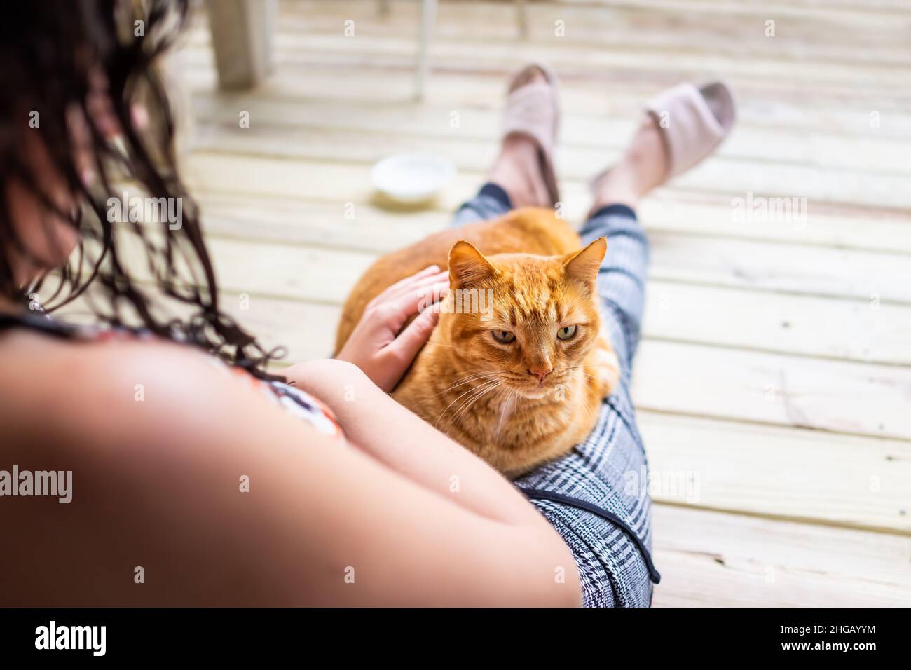 Young woman sitting on floor holding in arms lap cat lying down petting stroking feline orange ginger kitty outside at home house balcony porch patio Stock Photo