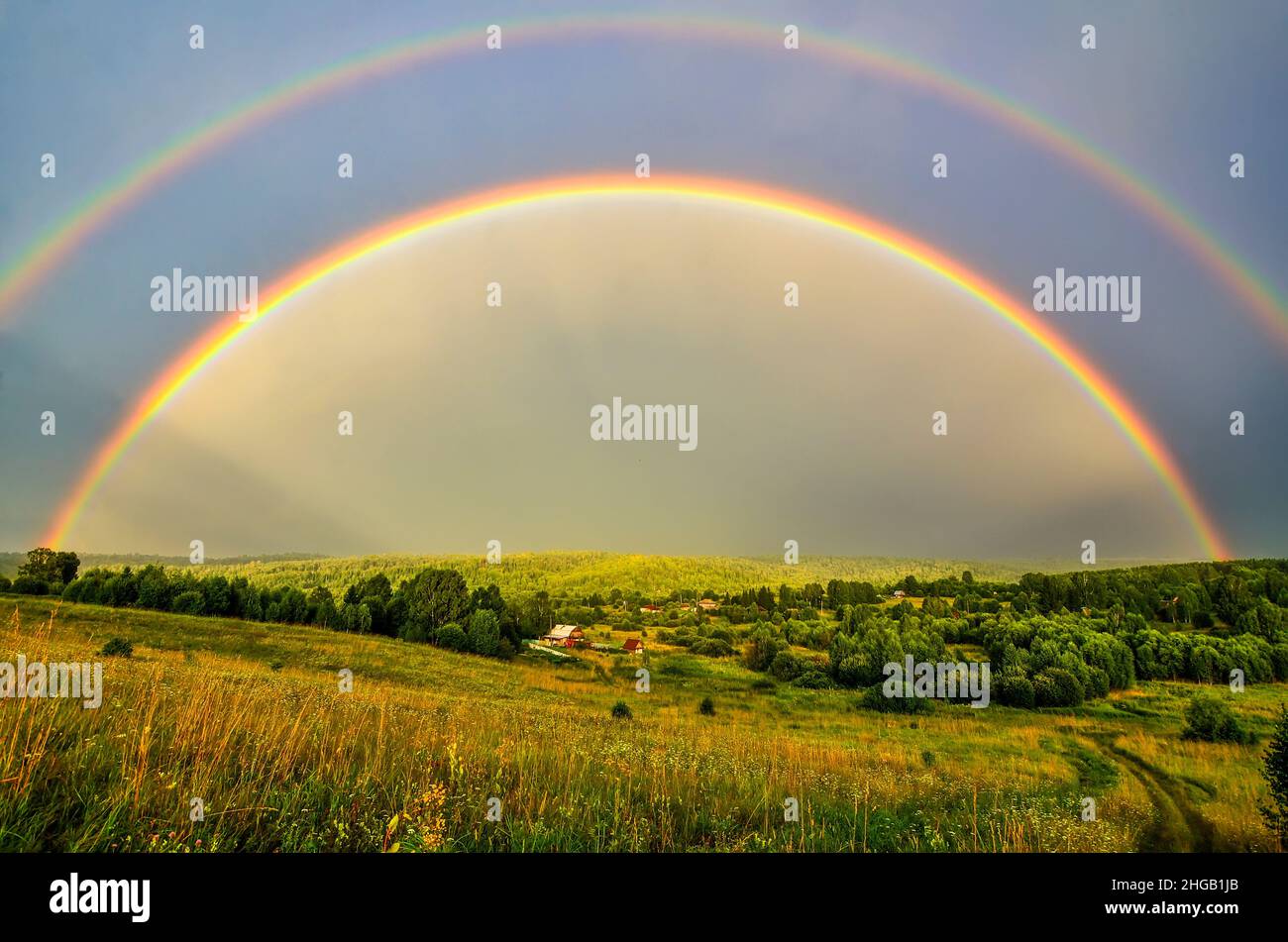 Full arc of double rainbow over summer evening rural landscape - panoramic view of hills with deep forests covered, golden colored meadow with sun bea Stock Photo