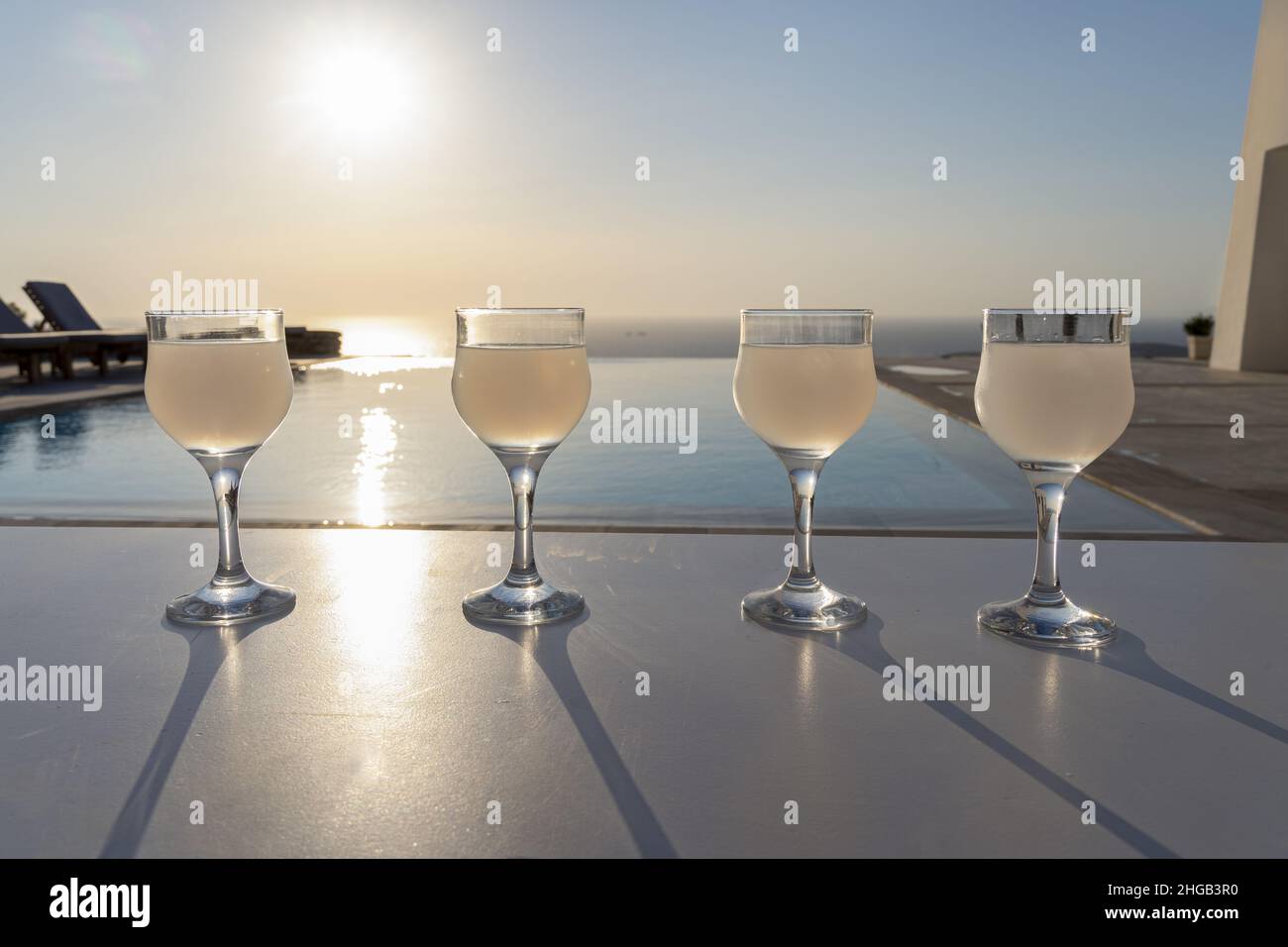Close up of four ouzo stemmed glasses on a white stand during sunset with the Mediterranean sea in the background Stock Photo
