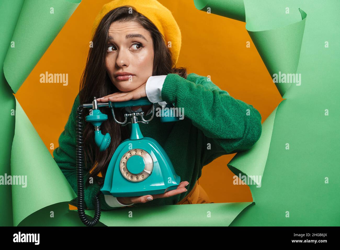 Young sad white woman peeking out hole isolated on green background holding landline phone Stock Photo