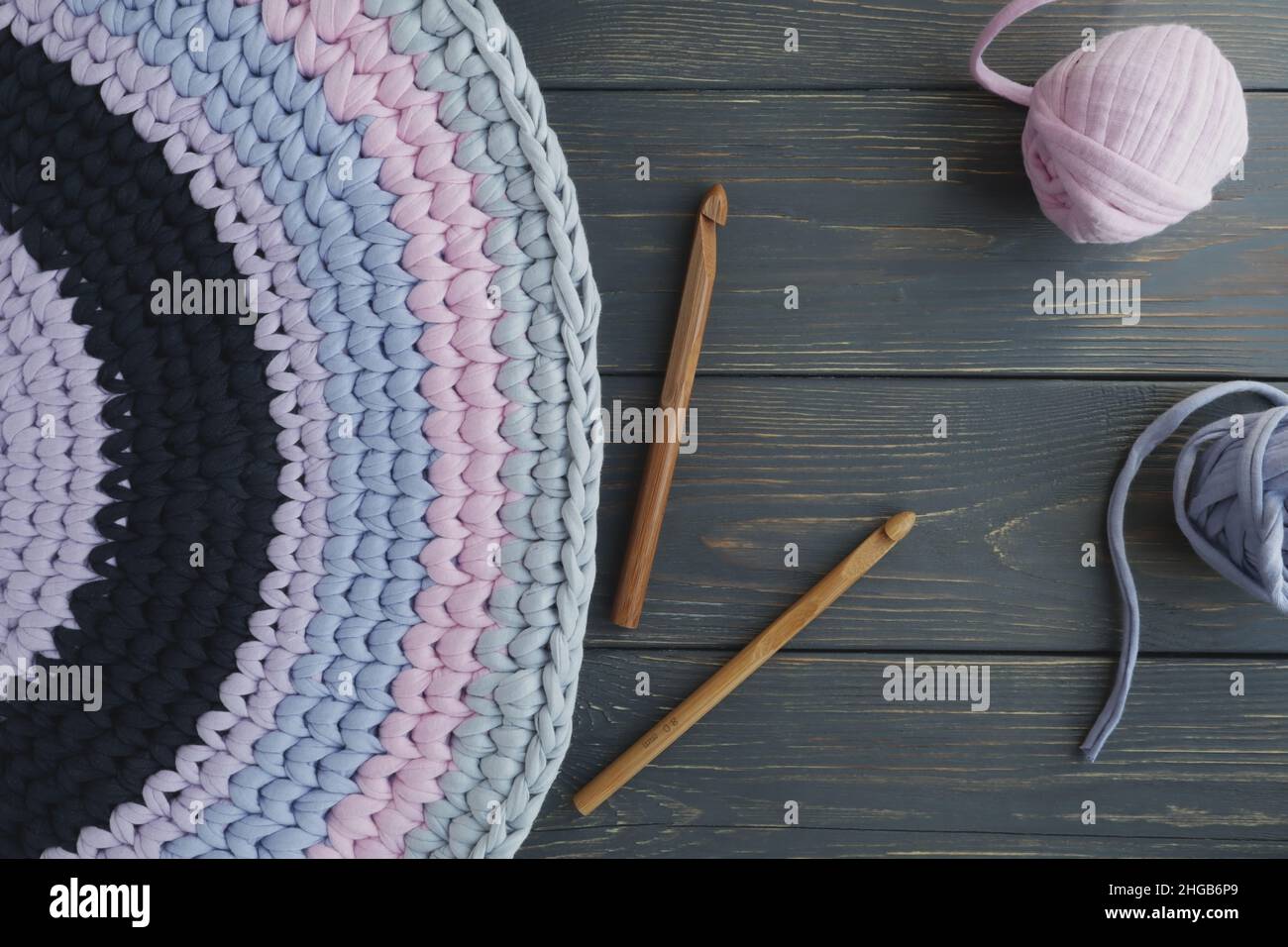 A mat made of knitted yarn and a crochet hook on a gray wooden background with space for text. Home needlework. Stock Photo