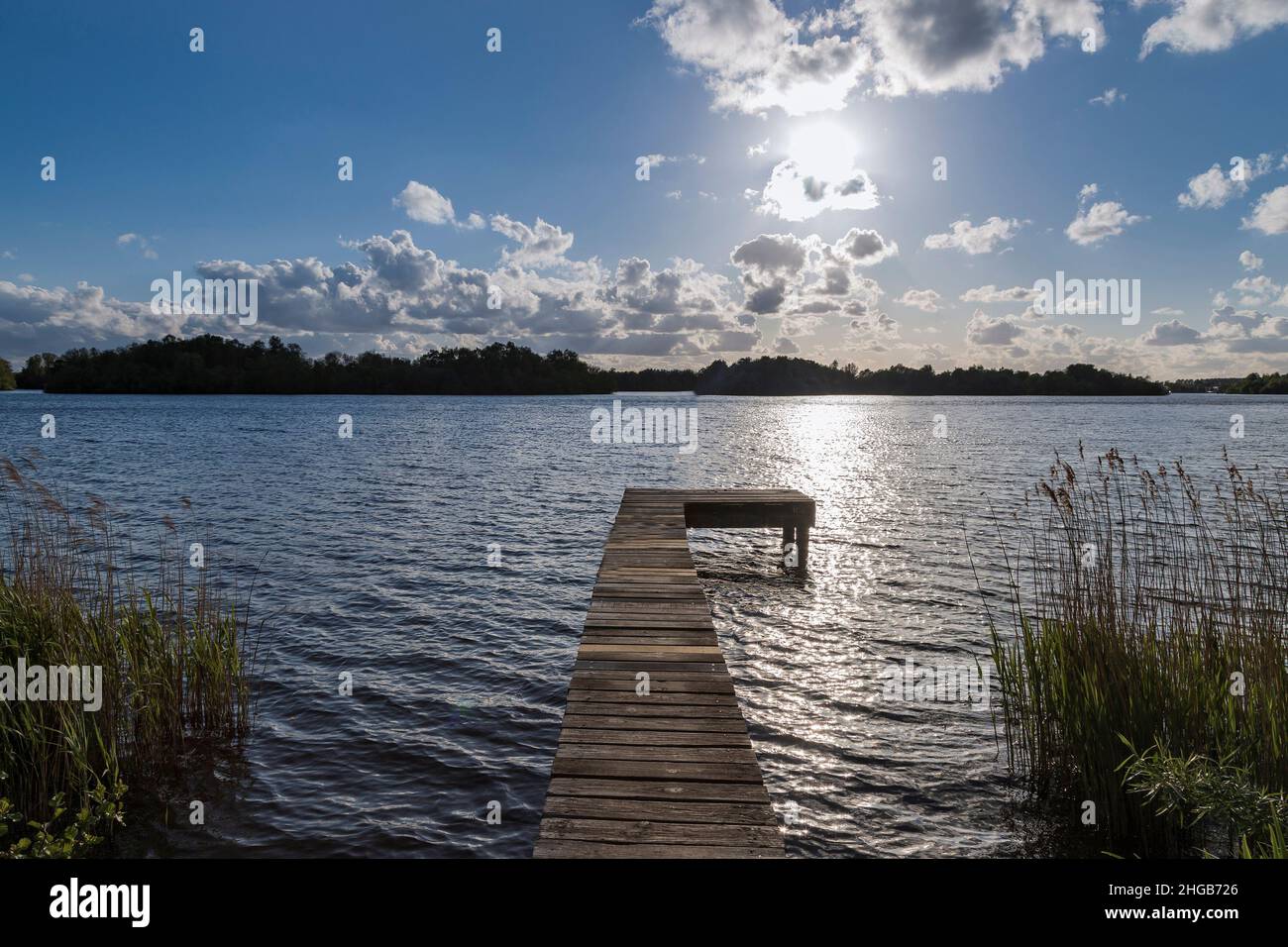 Water canal at Glimmen site in Holland. The background is a blue sky with white clouds. A wooden pier leads into the water. Stock Photo