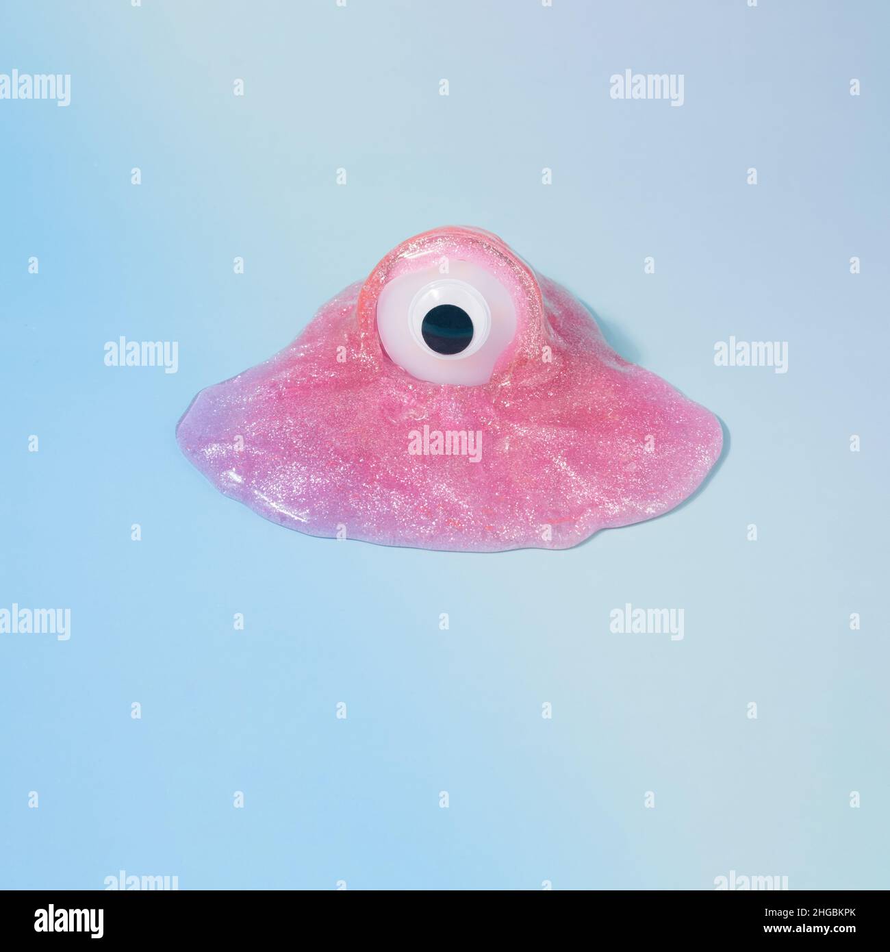 Monster figurine with one eye in a pink glitter slime on a gradient blue background. Flu virus funny concept. Stock Photo