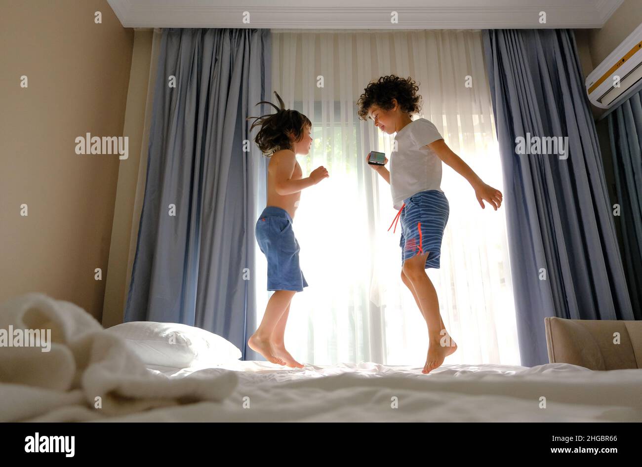 Two happy kids jumping on the bed with a phone listening to music Stock Photo