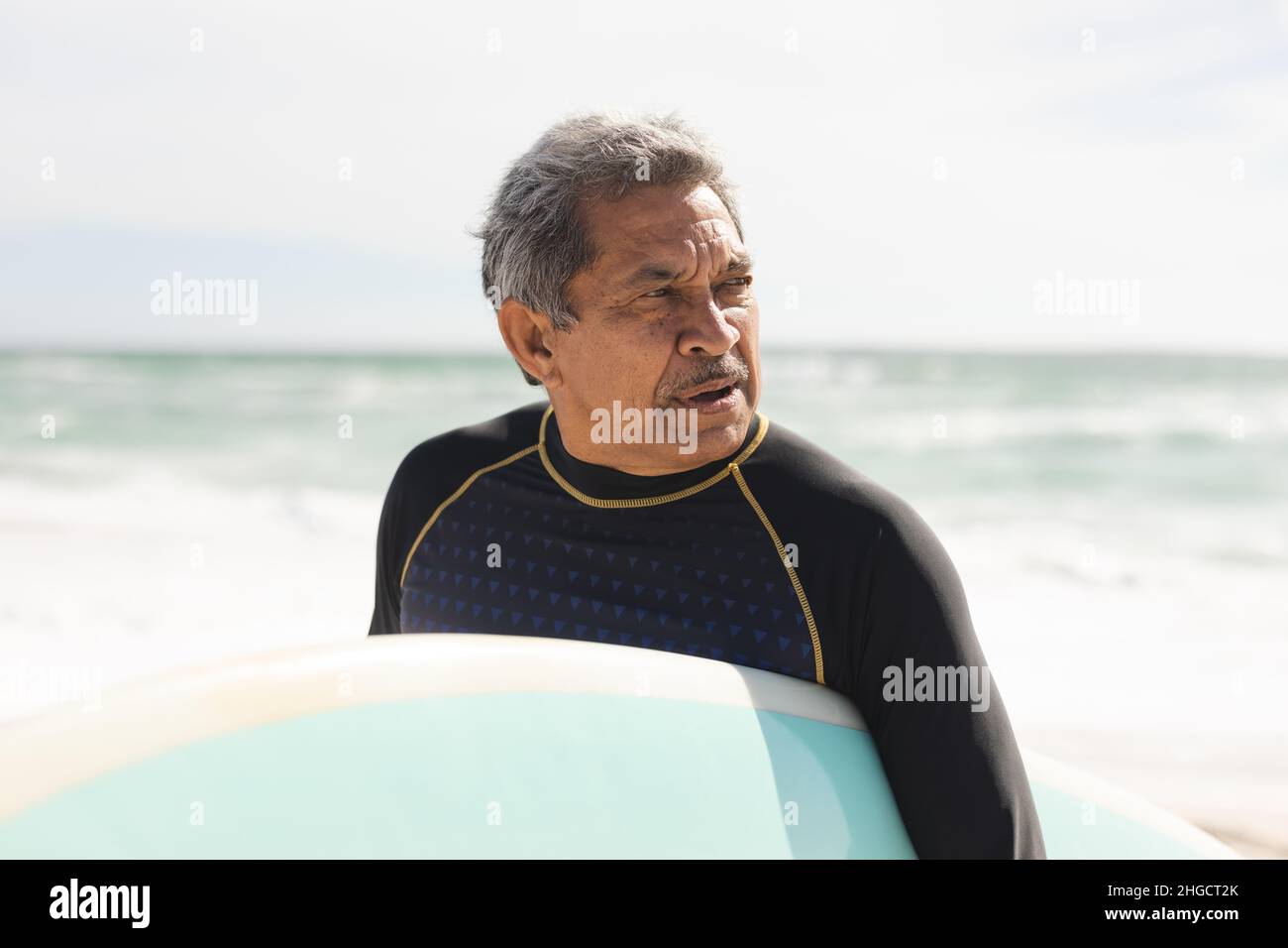 Retired biracial senior man looking away holding surfboard at beach against sky during sunny day Stock Photo