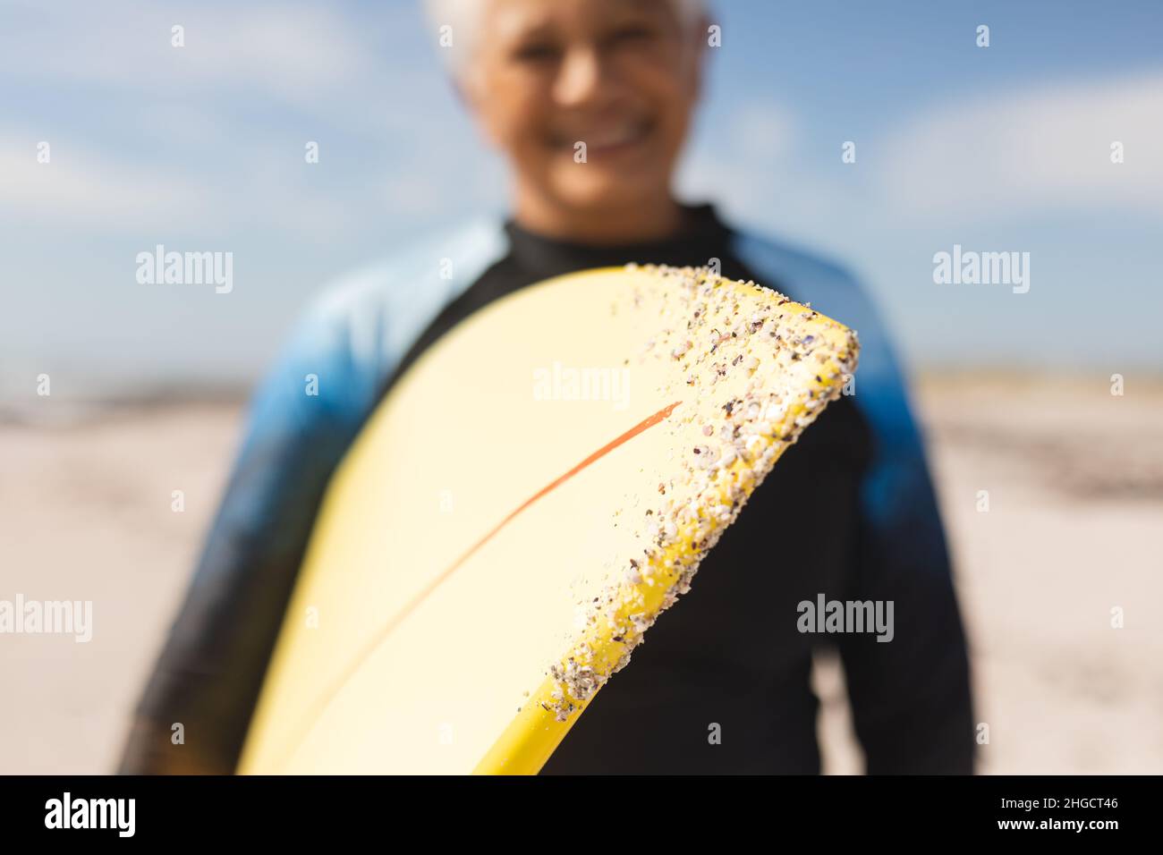 Surfboard with sand held by smiling biracial senior woman at beach during sunny day Stock Photo