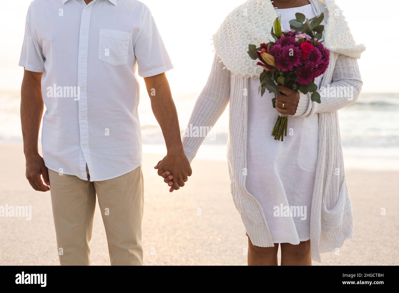 Midsection of biracial man holding hands with woman holding flower bouquet at beach wedding ceremony Stock Photo