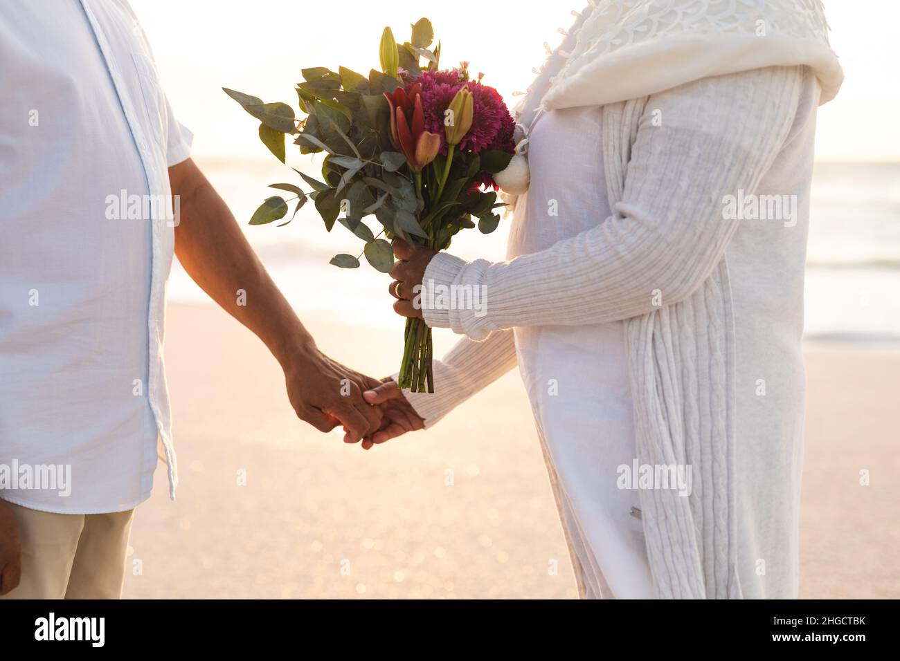 Midsection of multiracial senior couple holding hands with bouquet during wedding ceremony at beach Stock Photo
