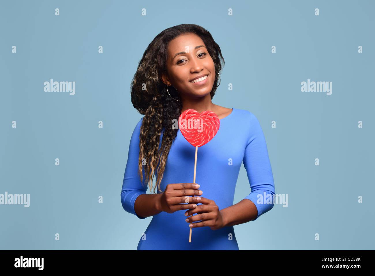 Beautiful black girl in a blue dress smiles and holds a large heart-shaped lollipop on a stick. Stock Photo