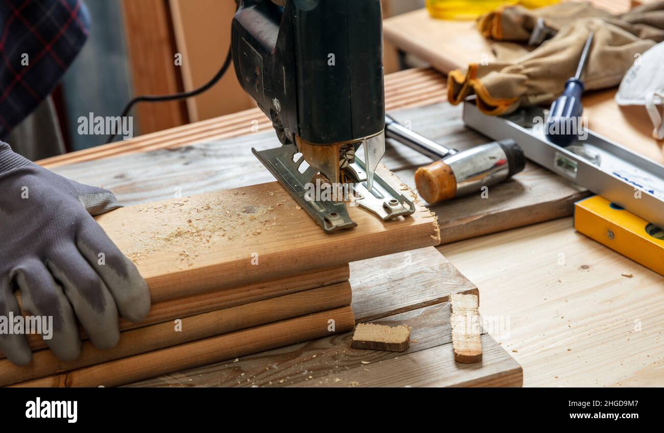 Electric jigsaw, carpenter gloved hand cutting wood with an electrical saw. Construction industry, work bench table closeup view Stock Photo