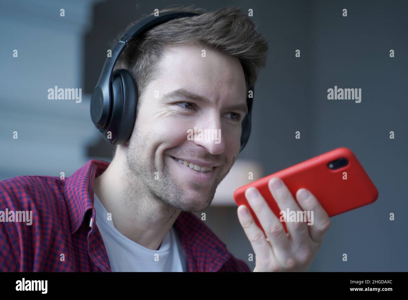 Cheerful young german man in wireless headphones holding smartphone in hand, recording audio message Stock Photo