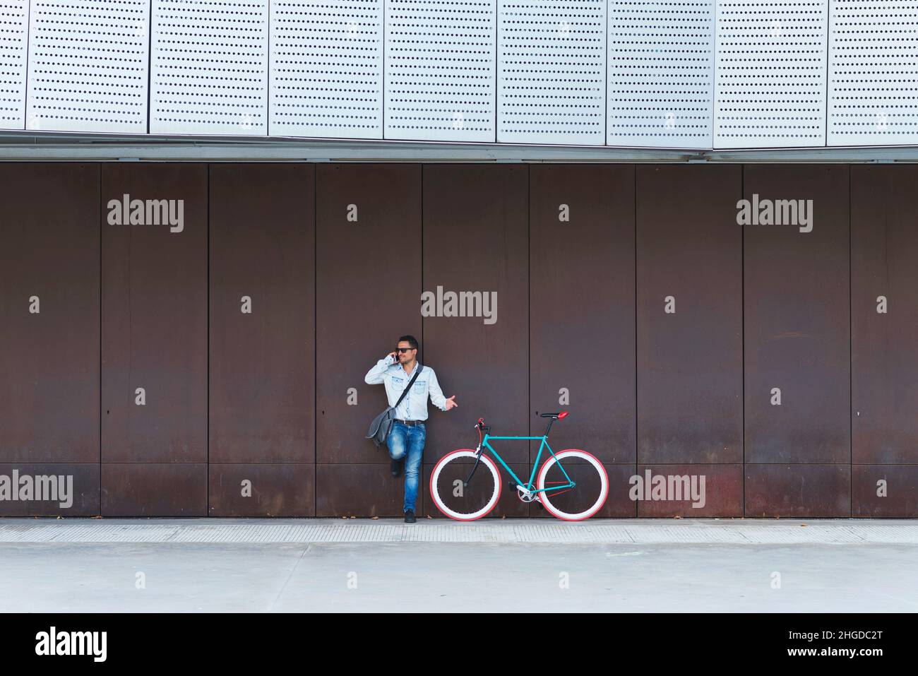 Young male with sunglasses and shoulder bag standing nest to a fixed gear bike while calling by phone Stock Photo