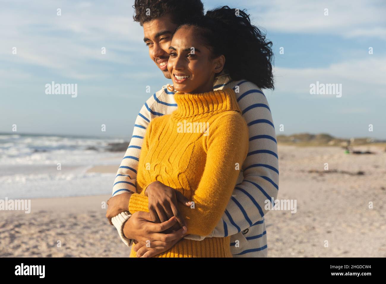 Happy multiracial couple embracing each other while looking away at beach against sky Stock Photo