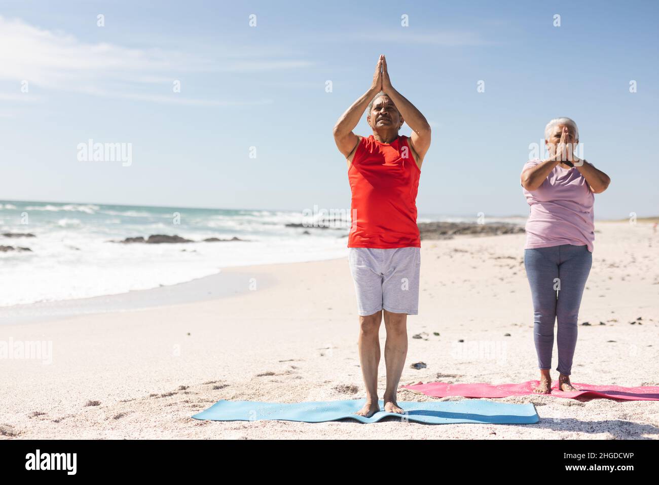 Full length of biracial senior couple with hands clasped practicing yoga asana at sunny beach Stock Photo