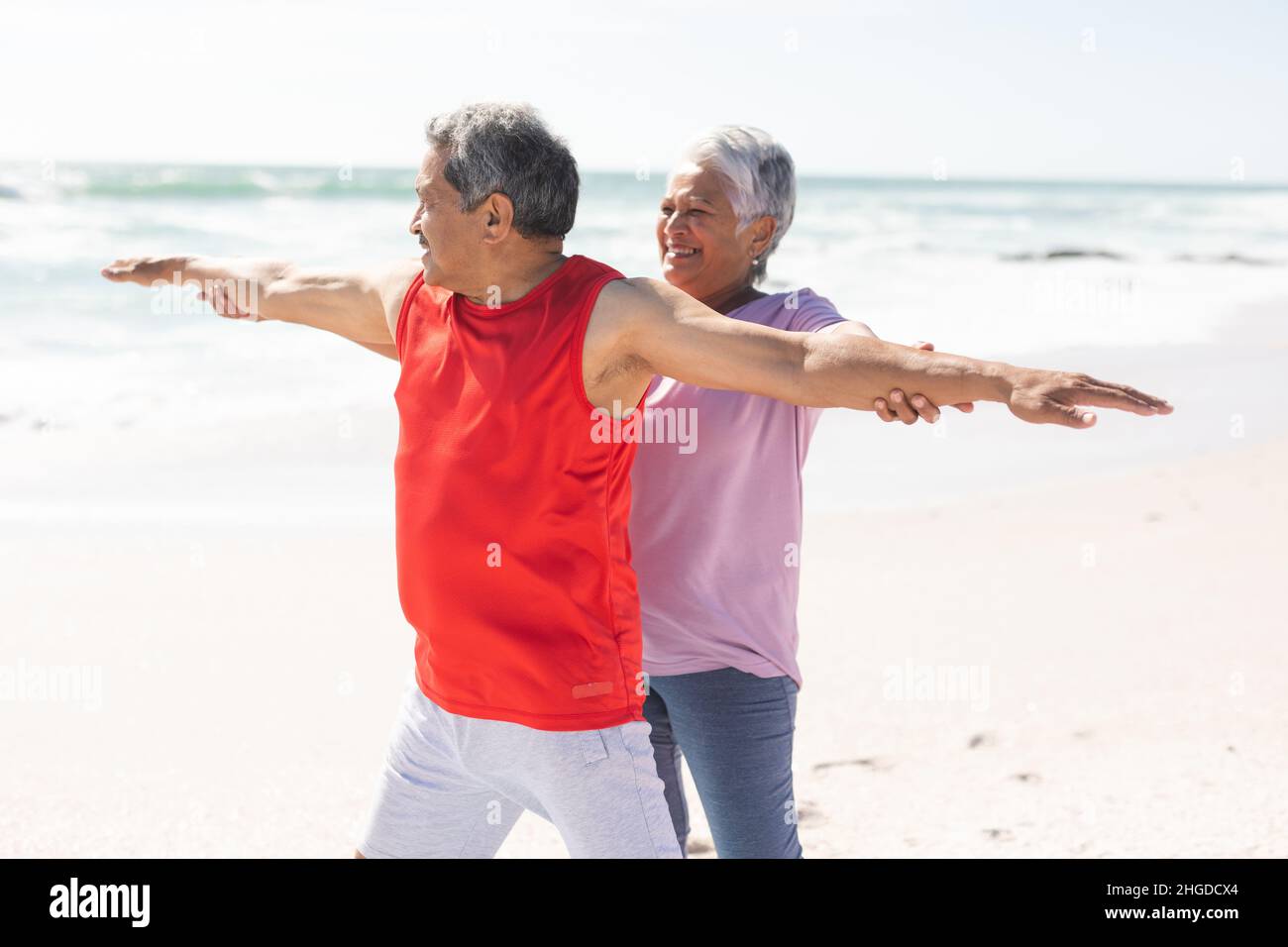 Smiling senior biracial woman helping man with arms outstretched yoga posture at sunny beach Stock Photo