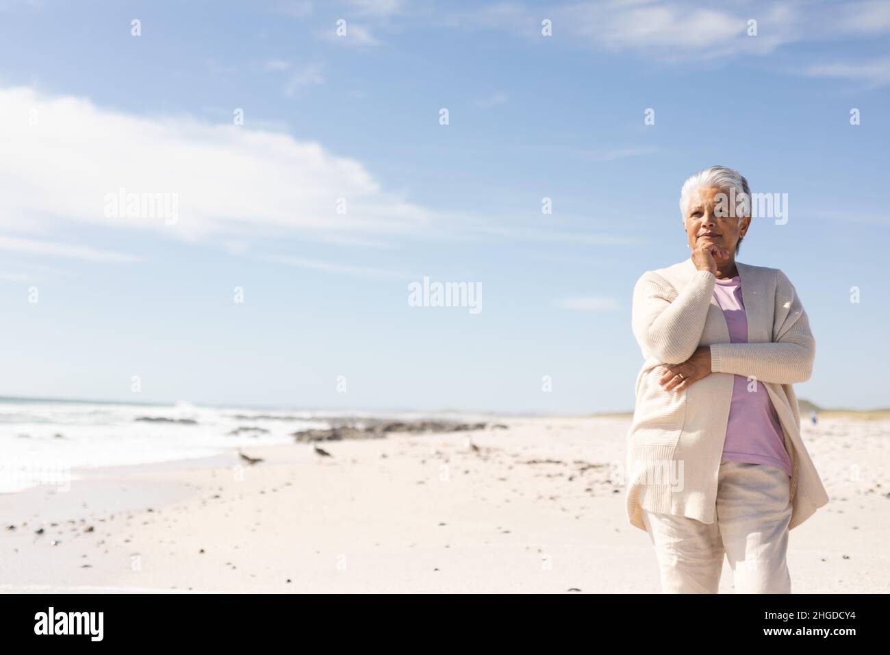 Retired biracial senior woman with hand on chin contemplating at beach against sky during sunny day Stock Photo
