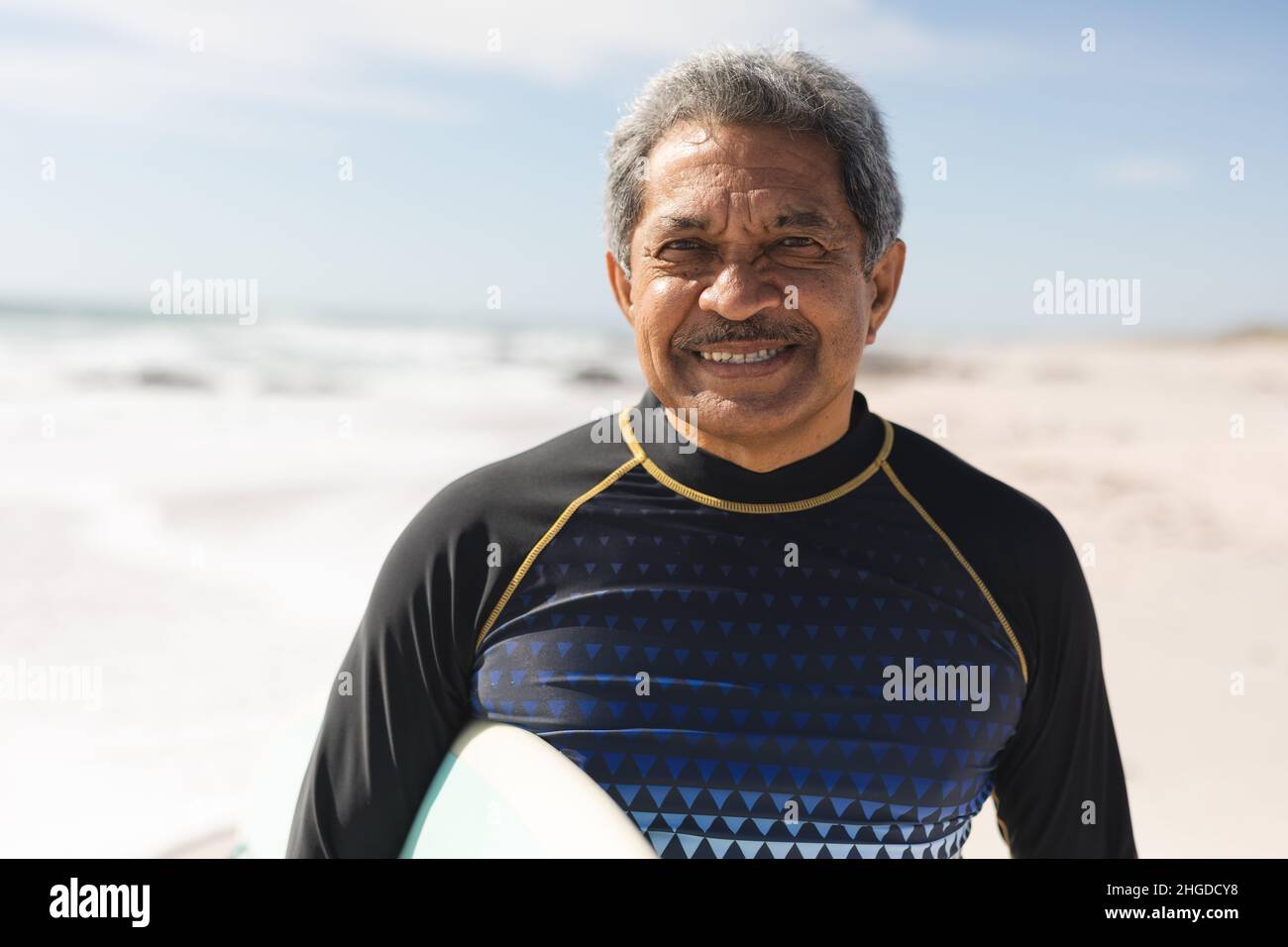 Portrait of smiling retired biracial senior man standing with surfboard at beach on sunny day Stock Photo