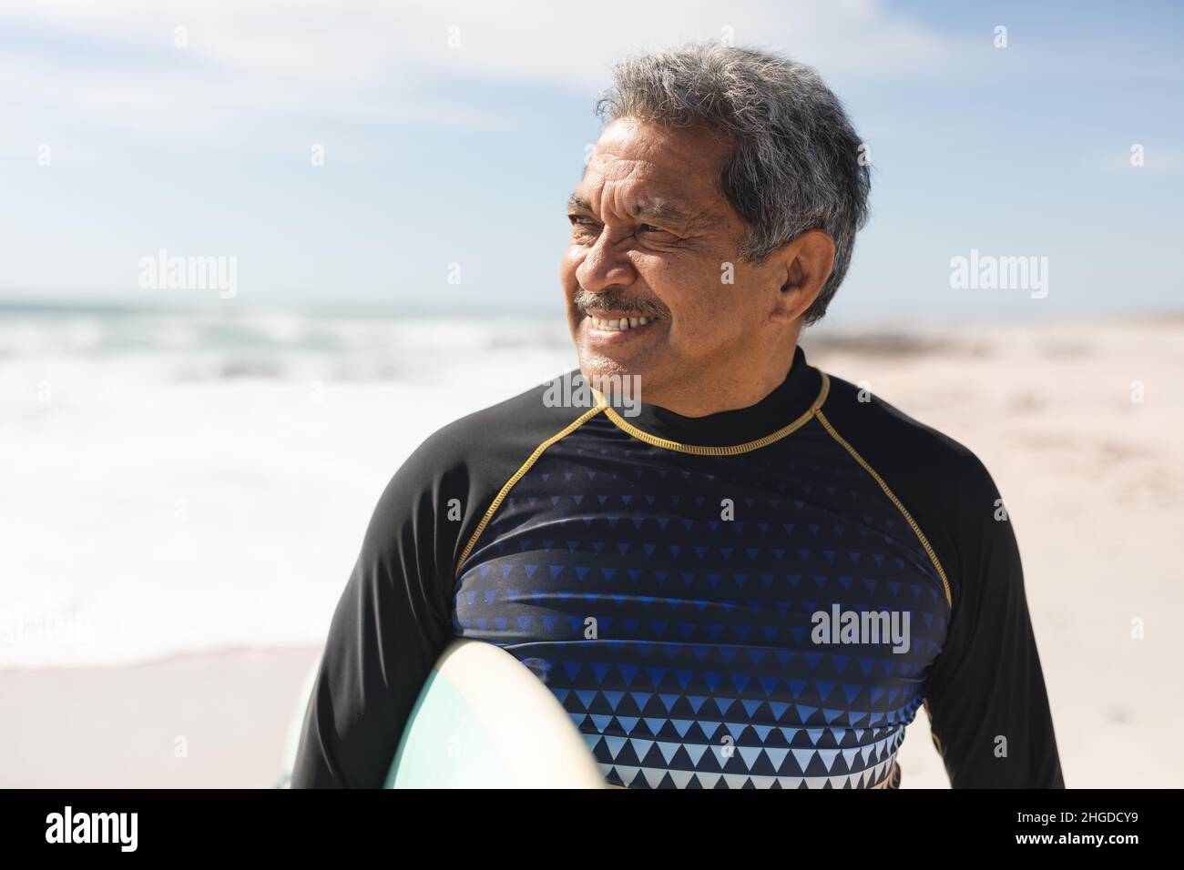 Smiling retired biracial senior man looking away while standing with surfboard at beach on sunny day Stock Photo