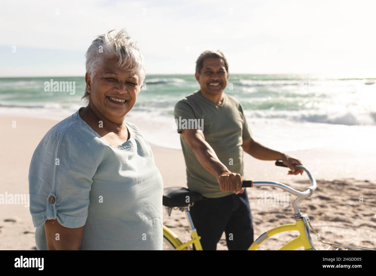 Portrait of smiling retired senior multiracial couple wheeling bicycle at beach on sunny day Stock Photo