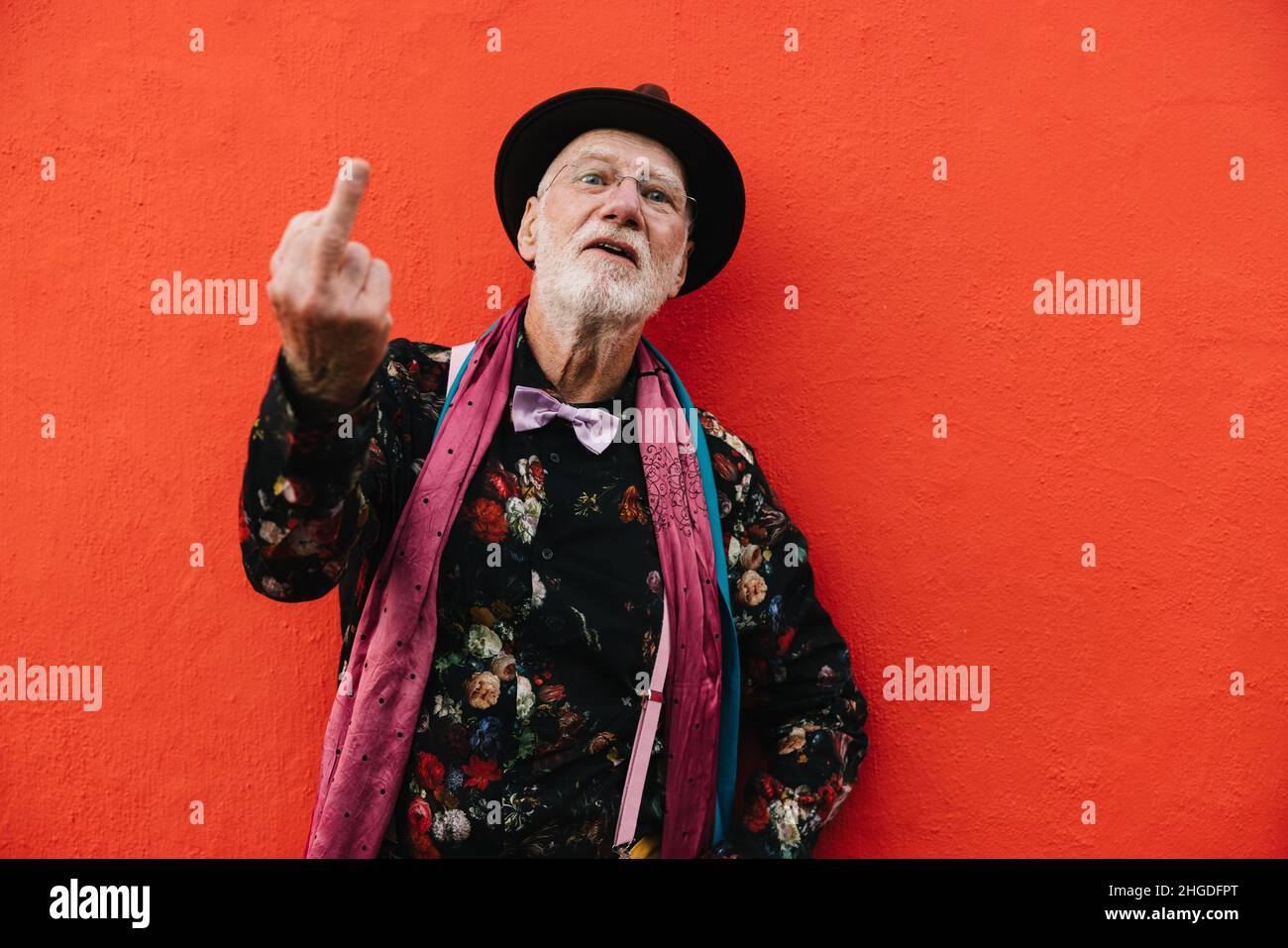 Mature man showing his middle finger against a red background. Senior man looking at the camera while making a swear gesture. Retired elderly man wear Stock Photo