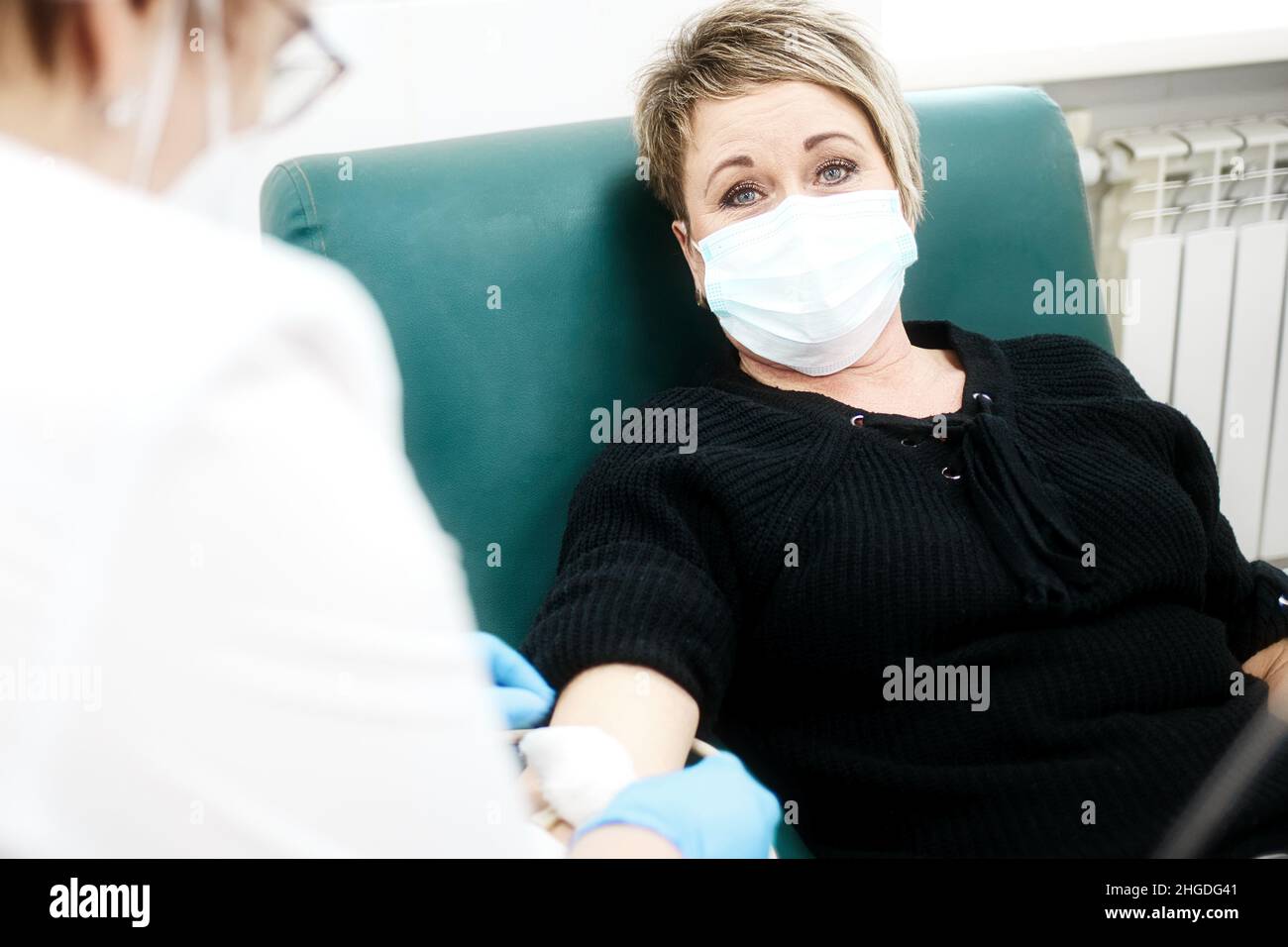 Rehabilitation and treatment in medical clinic. 40-50 year old woman in medical mask sits in chair and looks into camera. Nurse injects woman into vein. Stock Photo