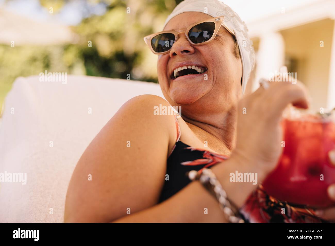 Senior woman enjoying a tiki cocktail while vacationing in the summer. Carefree elderly woman laughing cheerfully while relaxing on a lounger. Happy m Stock Photo