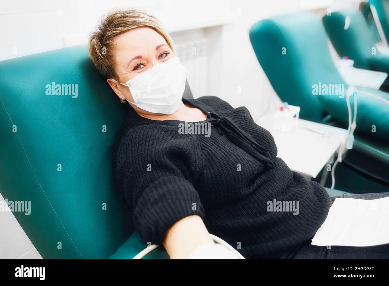 Woman of 40-50 years old in medical mask on her face sits in chair and donates blood from vein. Donation and care for sick. Real scene. Stock Photo