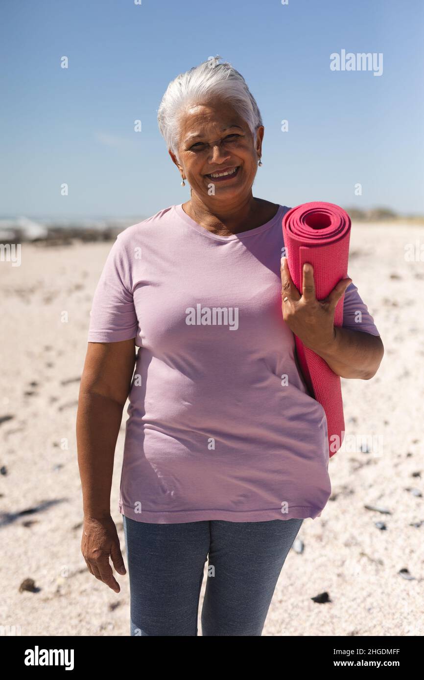 Portrait of smiling retired senior biracial woman holding yoga mat with short white hair at beach Stock Photo