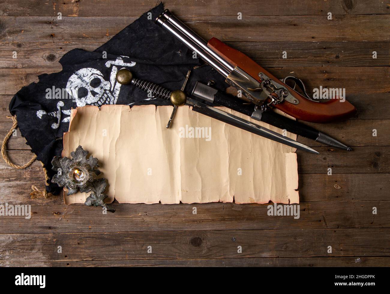crossed vintage flintlock pistol and large pirate dagger and jolly roger flag and blank papyrus with space for text or card on rough wooden table Stock Photo