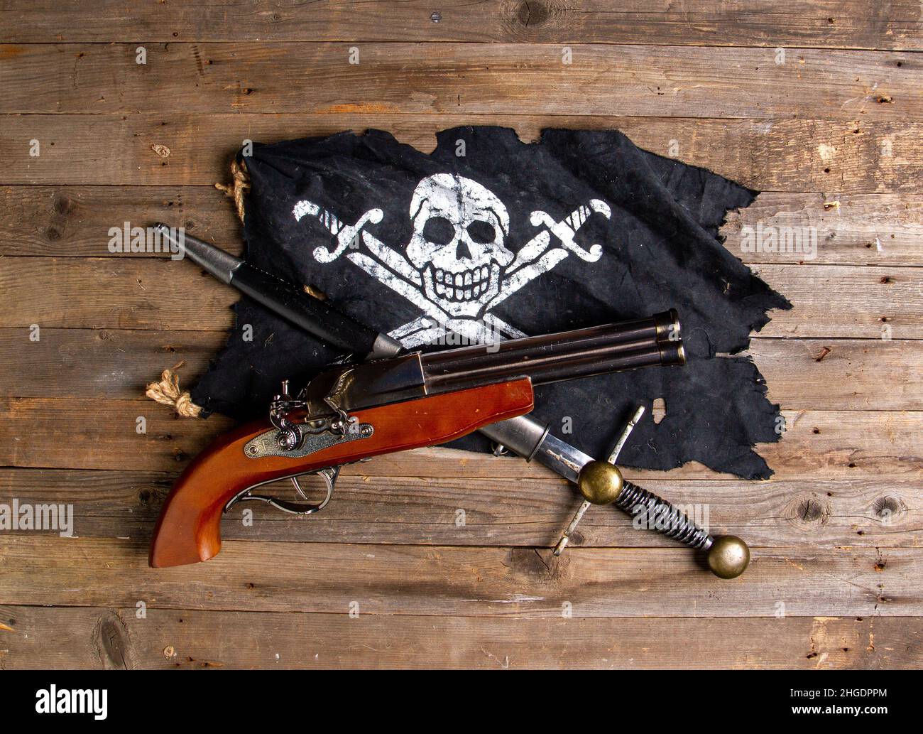 crossed antique flintlock pistol and large pirate dagger and jolly roger flag on a rough wooden table Stock Photo