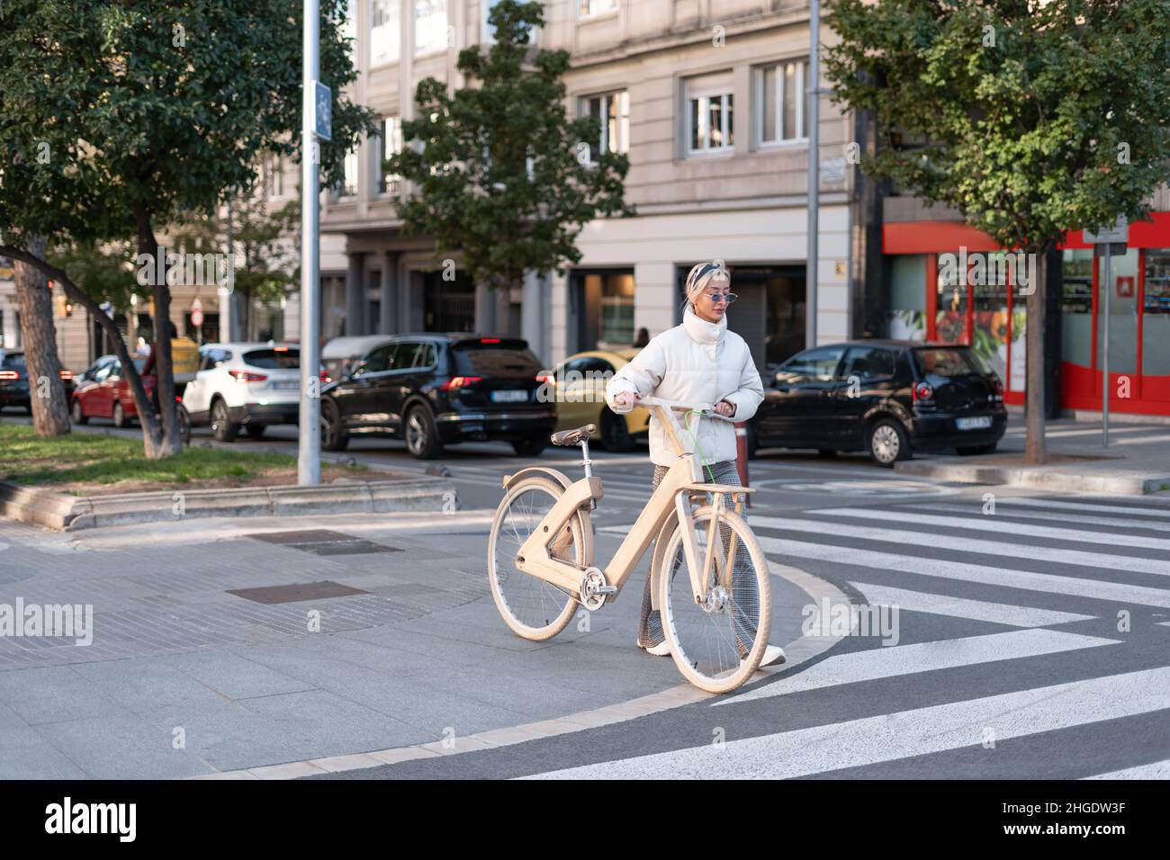 Blond woman with lumber eco friendly bicycle walking on sidewalk near crosswalk on street of contemporary city. Stock Photo