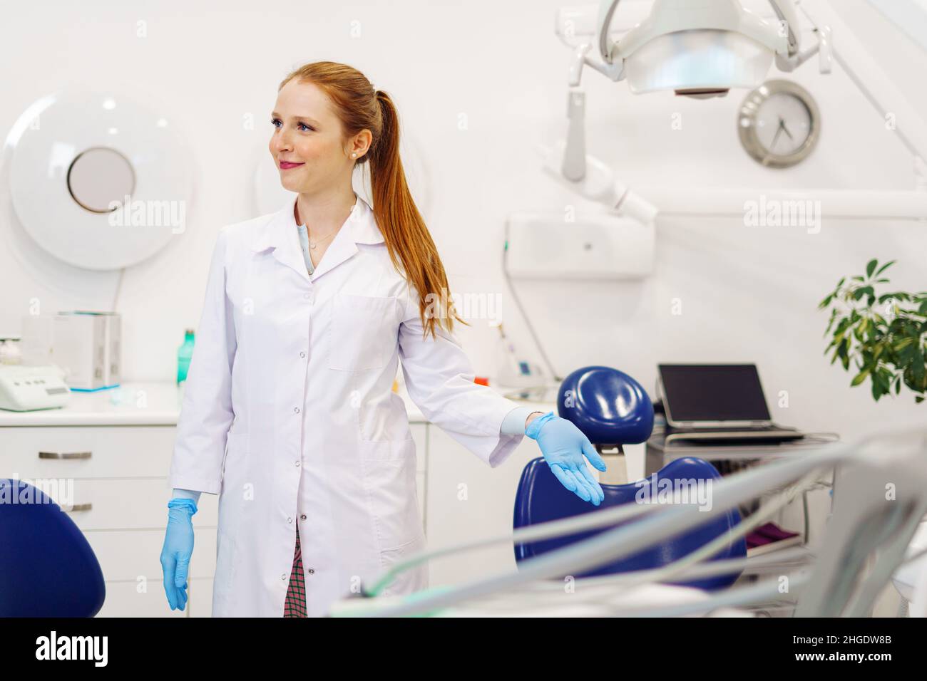 Glad woman in medical uniform with ginger ponytail smiling and showing empty chair while inviting patient to take seat in modern dentist office Stock Photo