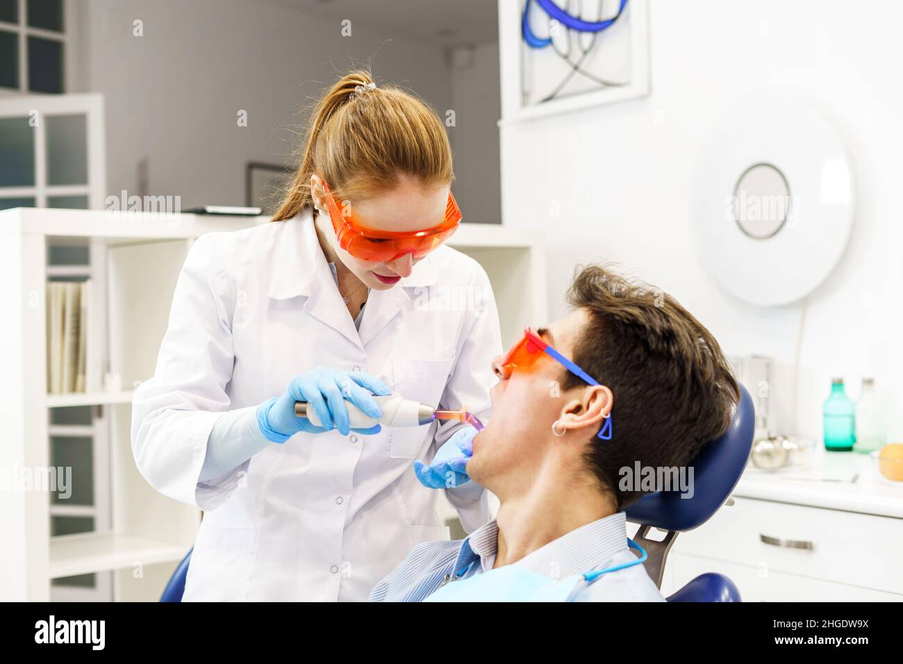 Female dentist in white robe with glasses and goggles using curing light on teeth of young man in office of contemporary dental clinic Stock Photo