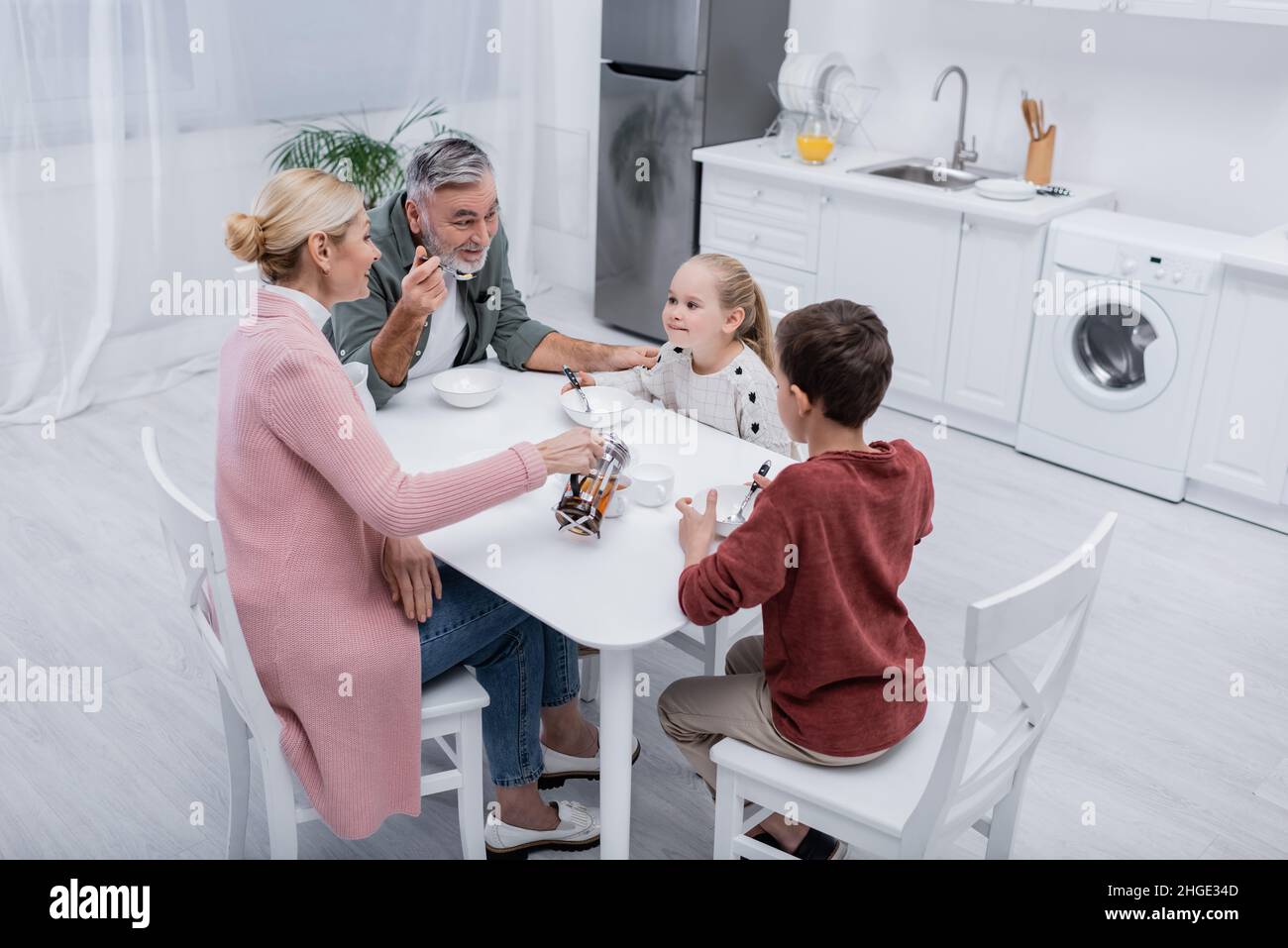 cheerful man talking to grandchildren near wife pouring tea during breakfast Stock Photo