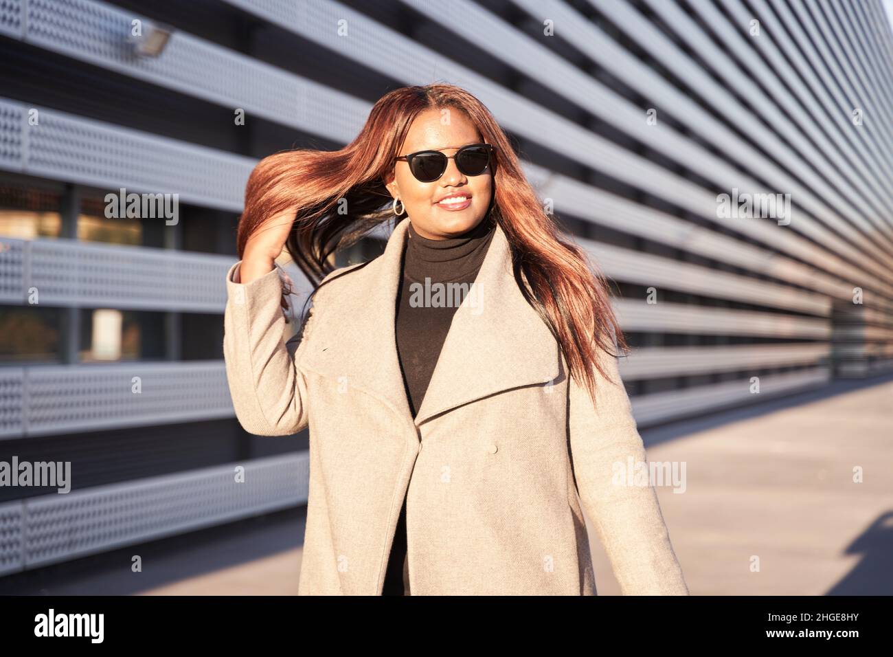 African-American woman wearing sunglasses. The successful young woman is wearing a business suit. She walks powerfully while combing her hair to one s Stock Photo